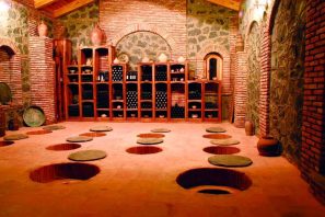 Adega subterrânea tradicional com paredes de tijolos e ânforas de barro enterradas no chão, usadas para fermentação e armazenamento de vinho, representando métodos antigos de vinificação. Geórgia – O Berço Perdido do Vinho Moderno