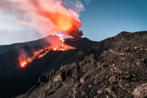 Vulcão em atividade liberando lava e fumaça sobre uma paisagem rochosa de origem vulcânica, ilustrando a formação dos solos minerais que, após milhares de anos de estabilização, dão origem a terroirs únicos para o cultivo de vinhos vulcânicos.