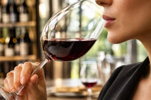 Pessoa segurando taça de vinho tinto próxima aos lábios em ambiente elegante de degustação, com fundo de adega iluminada e atmosfera de concentração sensorial. Vinho e Saliva: O Encontro Invisível que Transforma a Degustação.